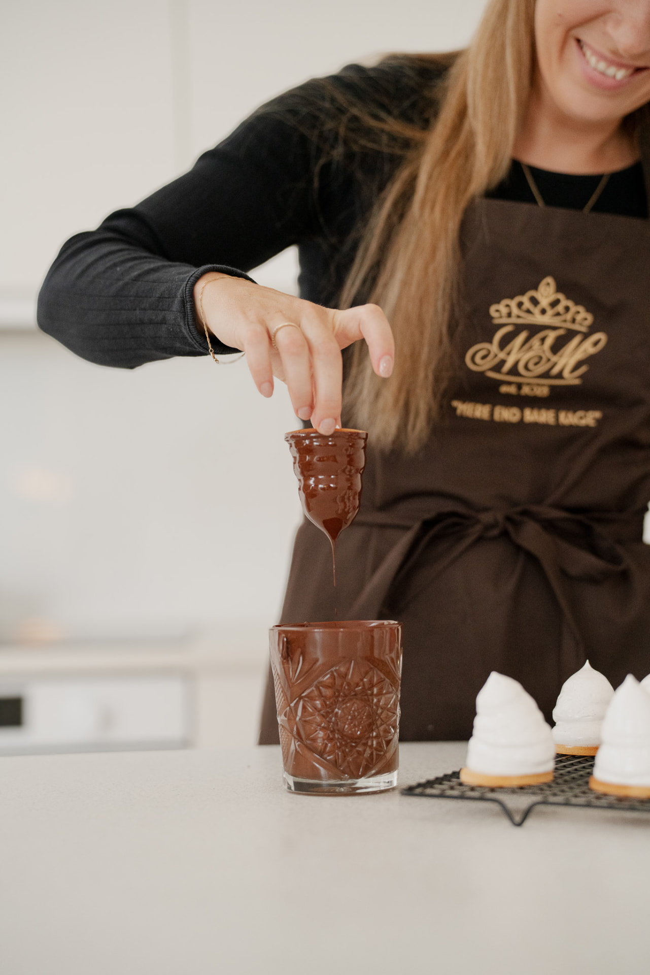 Nicole dypper en lille kage i et glas smeltet mørk chokolade iført brun forklæde